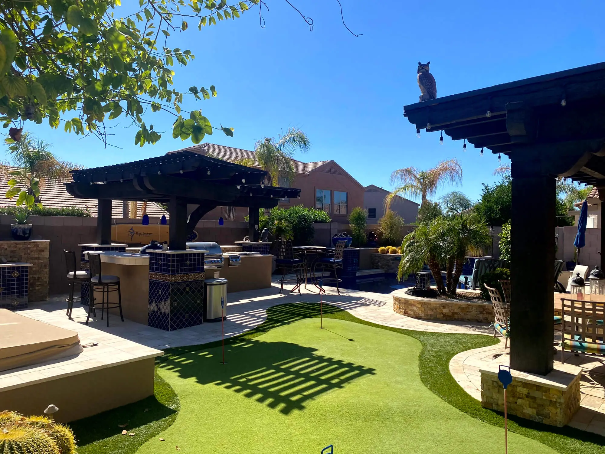 Upscale backyard oasis featuring a putting green and custom outdoor kitchen with dark-stained pergolas by Butchers Custom Painting.